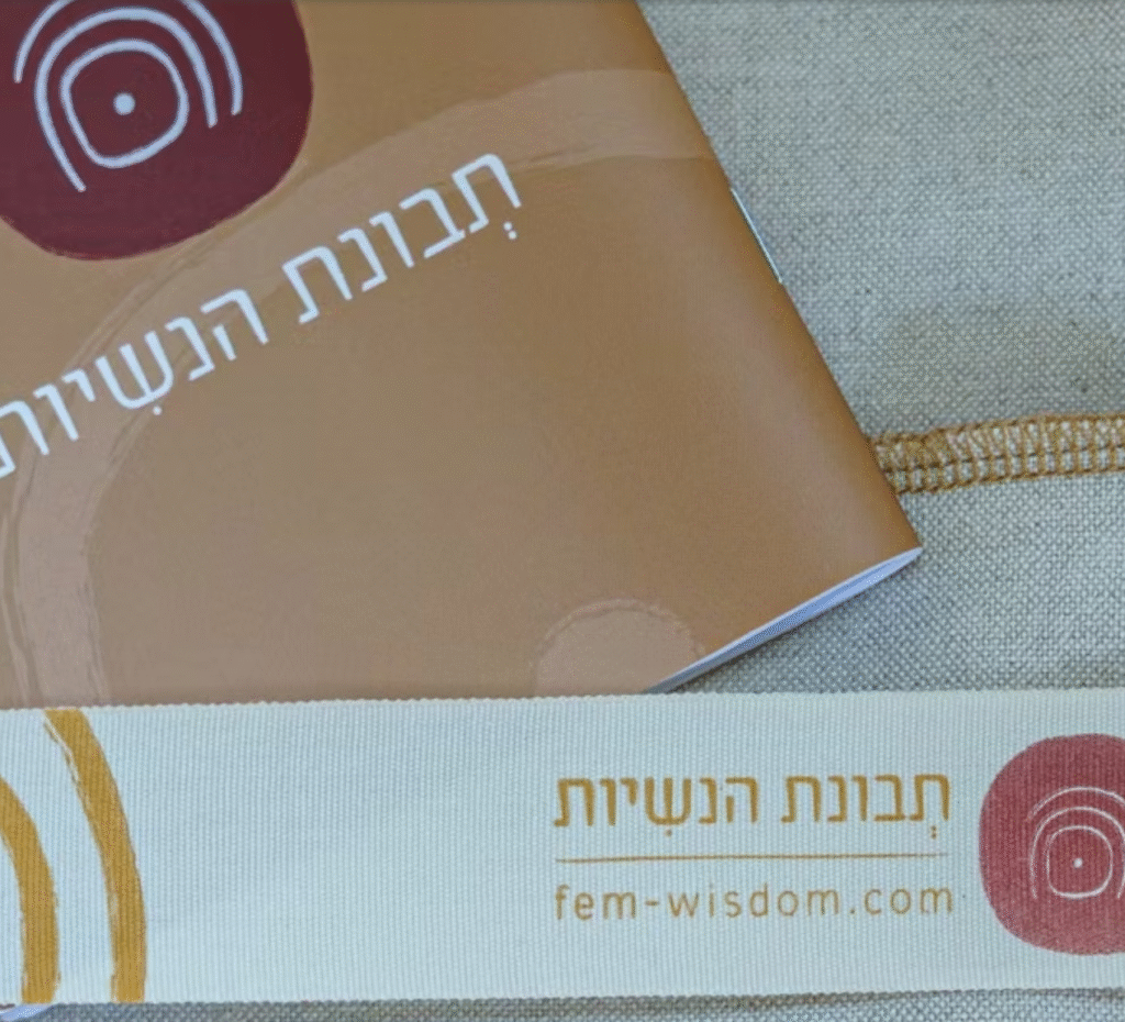 חוברת חומה ורצועת בד בצבע שמנת, שתיהן עם טקסט בעברית המתייחס ל"לידה שקטה" ול"חני גולד", מעוטרות בעיצובים מופשטים בצבעים אדום וצהוב. על הרצועה מופיע גם כתובת האתר fem-wisdom.com.