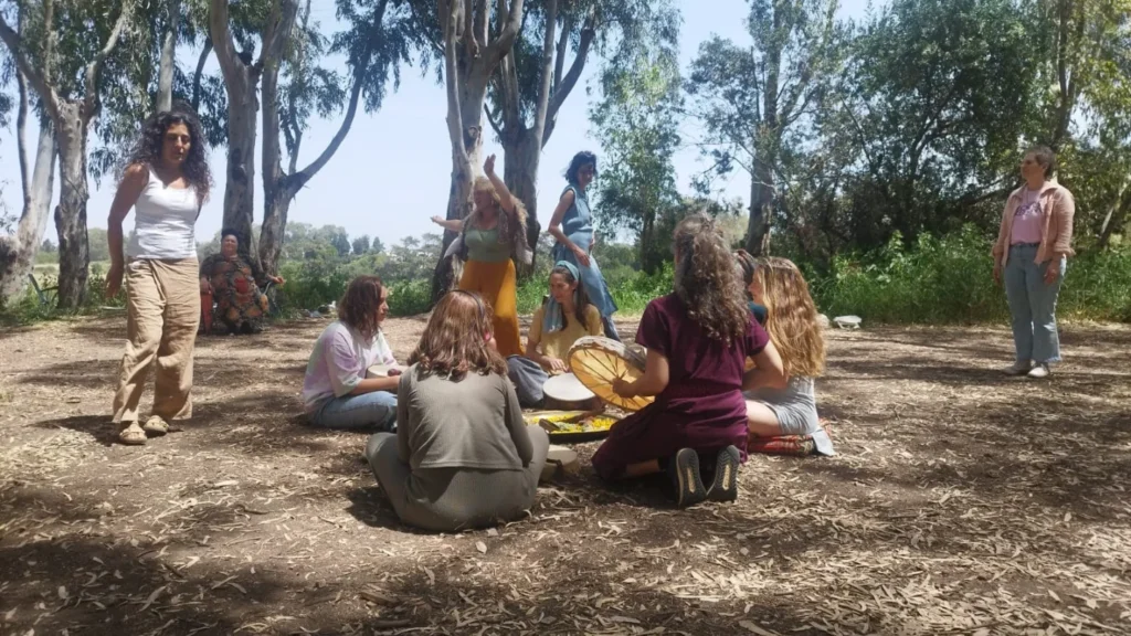 קבוצת אנשים, רובם נשים, יושבת במעגל על הקרקע בפינת יער שטופת שמש; חלקם אוחזים בכלי נגינה כמו תוף. האווירה הנינוחה והקהילתית מהדהדת בין העצים הגבוהים, על רקע שיחות שקטות בנושאים כגון הפלת הריון.