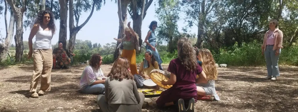 קבוצת נשים מתכנסת בחוץ, באזור מיוער ושטוף שמש. חלקן יושבות במעגל על הקרקע, ואחת מהן מנגנת בתוף. טקס מעגל נשי כחלק מתהליך עיבוד הפלה, לידה שקטה ולימודי הפדולה